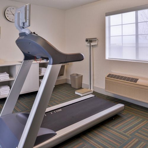 The image shows a small gym with a treadmill, scale, shelves with towels, a clock, a window, and an air conditioning unit.