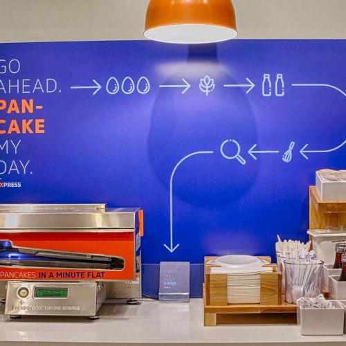 This image shows a pancake-making station with a conveyor oven, a sign saying 