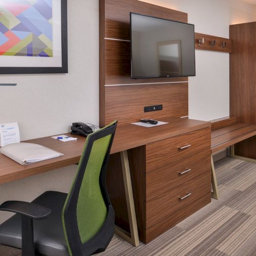 The image shows a hotel room with a desk, chair, TV, and wardrobe. There's a framed abstract artwork above the desk.