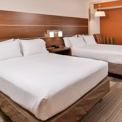 The image shows a hotel room with two large beds, white bedding, wood headboards, a striped chair, and a modern lamp on the wall.