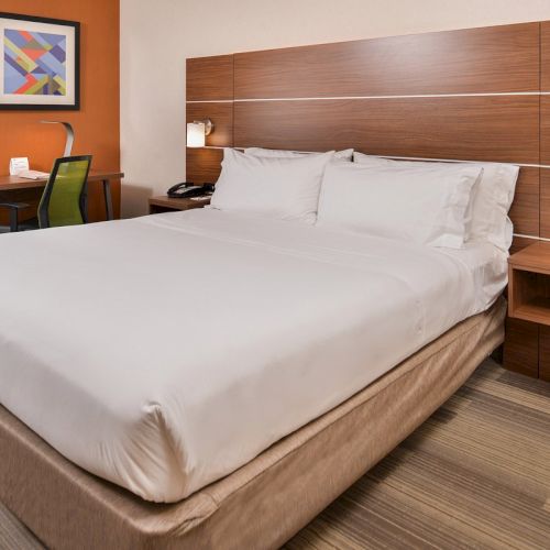 A neatly made bed in a modern hotel room, featuring a wooden headboard, two side lamps, and a desk with a chair and framed art.