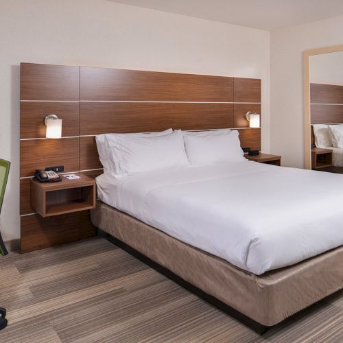 A modern hotel room with a large bed, wooden headboard, bedside lamps, desk with chair, and a mirror on the wall.