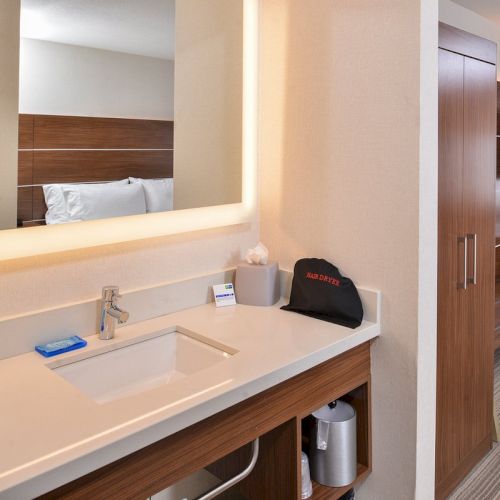 The image shows a hotel room with a bathroom vanity, towels, toiletries, a backlit mirror, TV, and wooden furniture, including a wardrobe.