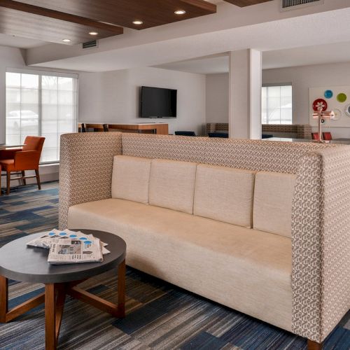 A modern lounge area with a beige sofa, round table with newspapers, orange chairs, wall art, and a TV. Bright, spacious, and inviting.