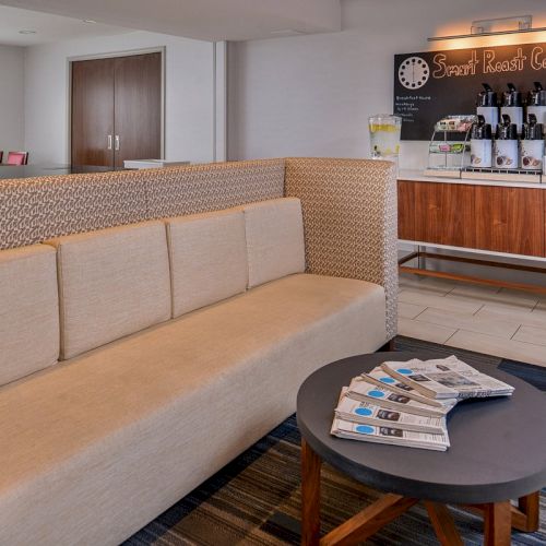 The image shows a cozy coffee area with a beige couch, a table with newspapers, and a coffee counter labeled 