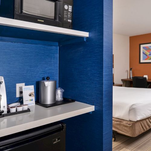 The image shows a hotel room with a small kitchenette featuring a microwave, coffee maker, and mini-fridge beside a bed and a desk area.