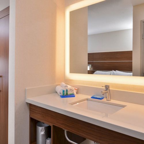 A hotel room bathroom with a backlit mirror, sink, toiletries, and a glimpse of the bedroom with a TV and seating area.