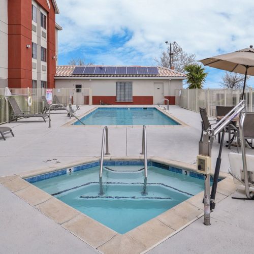 The image shows an outdoor pool area with a hot tub, lounge chairs, umbrellas, and a hotel building in the background.