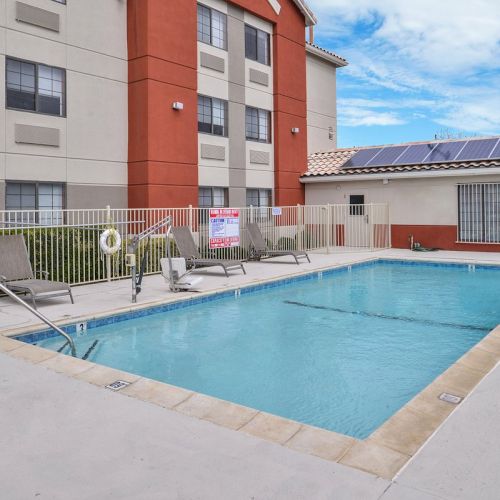 The image shows a small outdoor pool with lounge chairs, a safety fence, and a building with solar panels on the roof in the background.