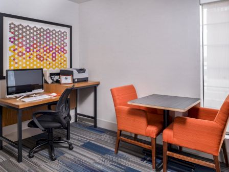 An office space with a desk, computer, printer, and two orange chairs around a small table. Framed artwork is on the wall.