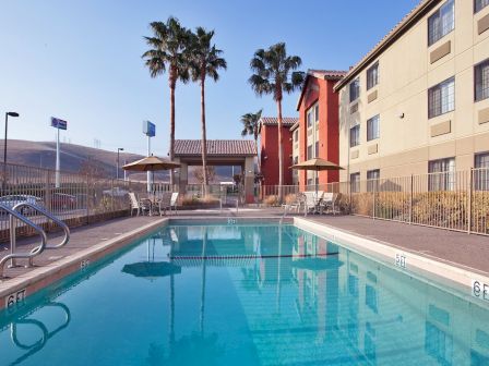 The image shows an outdoor swimming pool with clear water, surrounded by a fence, chairs, umbrellas, and palm trees next to a hotel building.