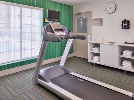 A gym room with a treadmill, white towels on shelves, a clock, and a large window letting in natural light completes the scene.