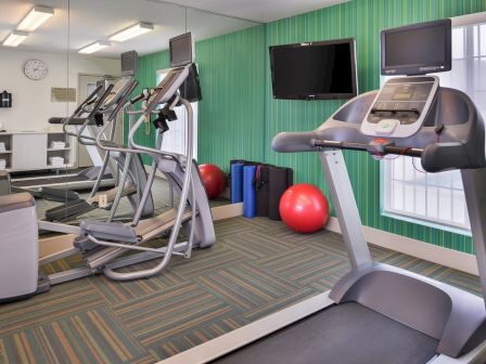 The image shows a gym with treadmills, elliptical machines, exercise balls, and wall-mounted TVs in a mirrored room with a clock.