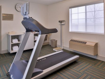 The image shows a small gym room featuring a treadmill, scale, towels on shelves, a wall clock, and a window.