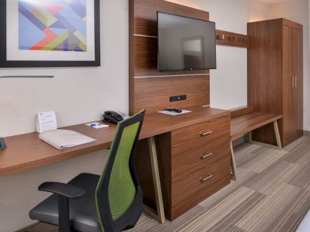 The image shows a hotel room with a desk, chair, TV, drawers, and artwork on the wall, featuring modern wooden furniture and decor.