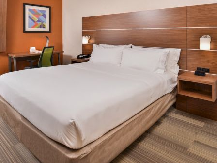 The image shows a modern hotel room with a neatly made bed, a wooden headboard, a desk with a chair, and a colorful framed artwork.