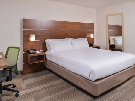 A hotel room with a double bed, wooden headboard, side tables, lamps, desk with chair, and a large mirror on the wall.