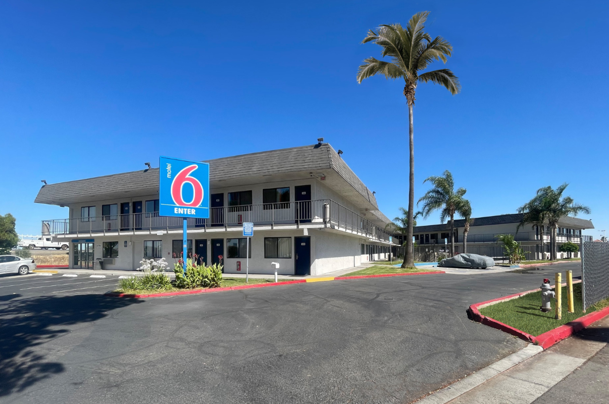 The image shows a Motel 6 building with a blue sign, surrounded by palm trees and a clear blue sky.