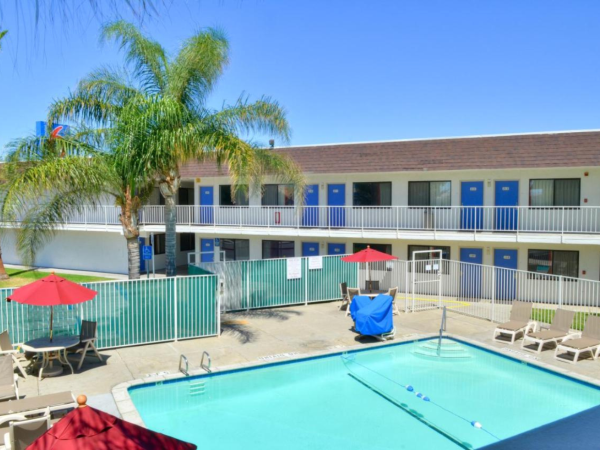 A motel with a blue and white exterior features a fenced outdoor pool, surrounded by red umbrellas and lounge chairs, under clear skies.