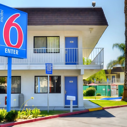 The image shows a Motel 6 building with a prominent sign, surrounded by palm trees and a parking area in the foreground.