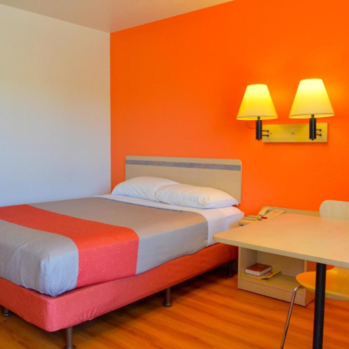 A hotel room with an orange accent wall, bed, side table with a chair, and two wall lamps.