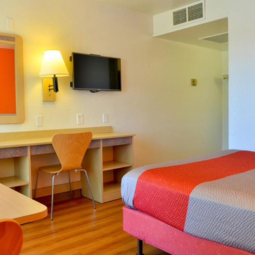 The image shows a hotel room with a bed, desk, chair, wall-mounted TV, and lamp, featuring orange and neutral tones.