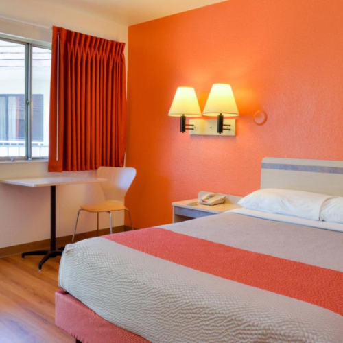 A brightly colored hotel room with a bed, wall lamps, a window with curtains, and a small table with a chair next to it.