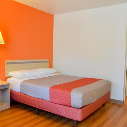 A simple hotel room features a bed, bedside table, and orange accent wall, with wooden flooring and a small open closet area.