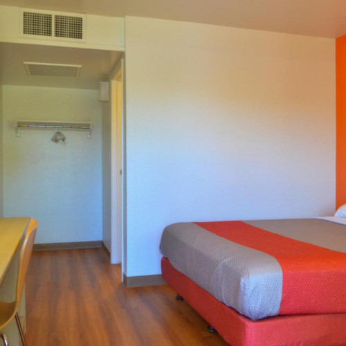 A simple hotel room with a bed, desk, lamp, and an orange accent wall. There's a small closet area with hangers near the entryway.