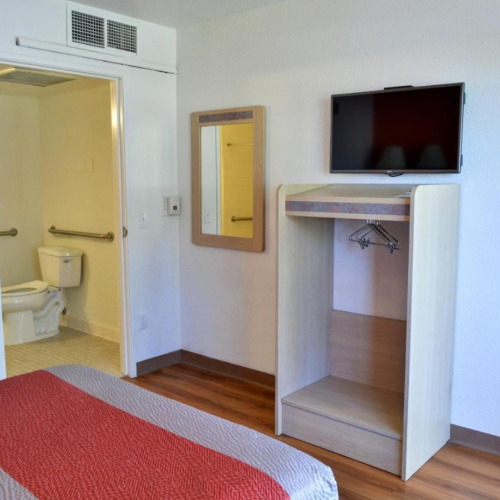 A hotel room with a bed, wall-mounted TV, open closet, and view of a bathroom with a toilet and grab bars.