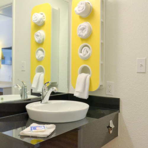 A bathroom with a sink, a mirror with rolled towels on a yellow rack, and a modern faucet.