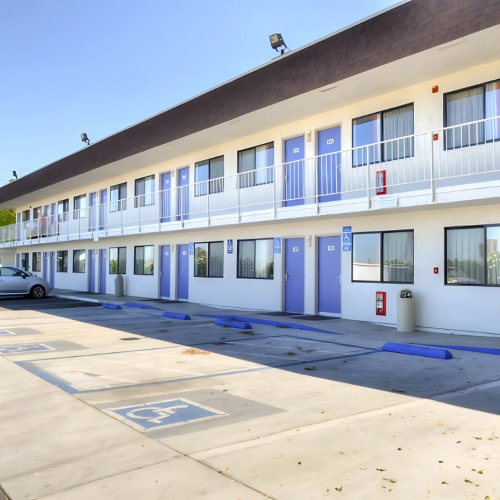 The image shows a two-story motel with a row of rooms, accessible parking spaces, and a few trees in the background under a clear sky.