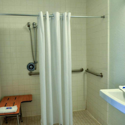 This image shows a bathroom with a shower curtain, a bench, handrails, a sink, and a mirror. It's designed for accessibility.