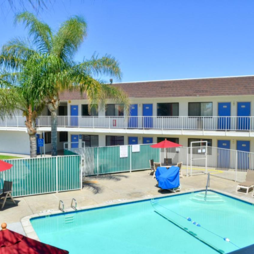 A motel courtyard with a swimming pool, surrounded by lounge chairs and red umbrellas. Palm trees and blue doors are visible in the background.