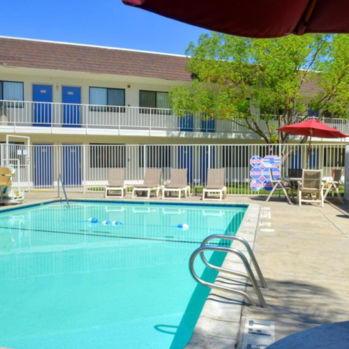 Outdoor pool area with lounge chairs, umbrellas, and a two-story motel in the background. Clear blue water and sunny weather.