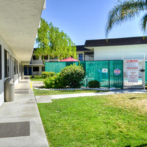 The image shows a motel exterior with rooms on the left, a grassy area, and a fenced pool in the background, surrounded by palm trees.