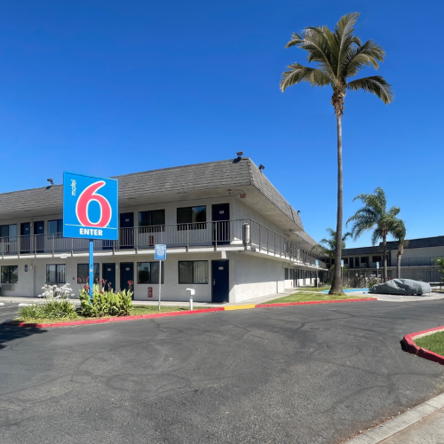 The image shows a Motel 6 building with palm trees and a large blue sign in a parking area under a clear blue sky.