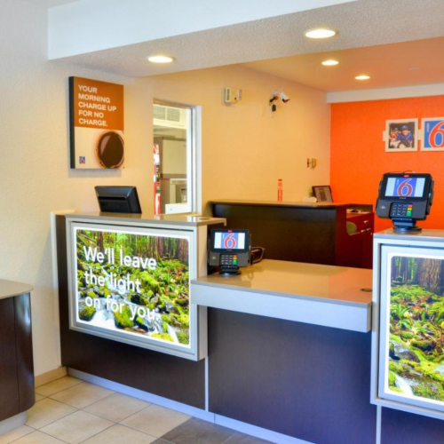 This image shows a brightly lit Motel 6 reception area with modern decor and promotional signs on the front desk.