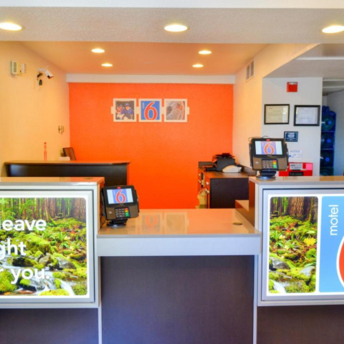 The image shows the reception area of a Motel 6 with a bright orange wall and signage saying 