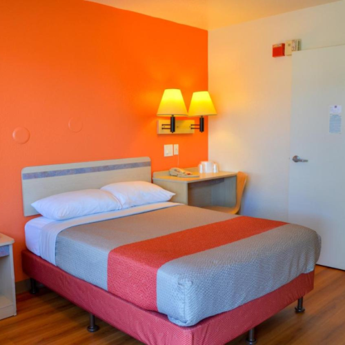 A simple hotel room with a bed, bedside tables, orange accent wall, lit lamps, wood flooring, and an open door to the bathroom.