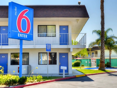 The image shows a Motel 6 with a blue and red sign, two-story building, and palm trees in the background, under a clear sky.