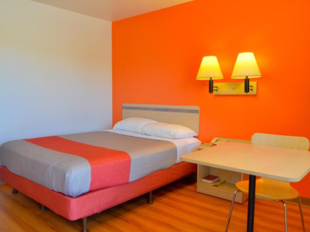 A brightly colored room with a bed, table, chair, and wall lamps against an orange accent wall.