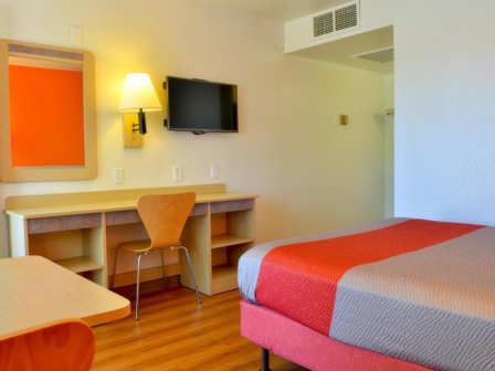 A hotel room with a bed, wall-mounted TV, desk, chair, lamp, and a mirror on the wall.