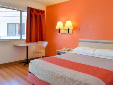 A hotel room with an orange accent wall, bed, side table, and a small desk with a chair near the window.