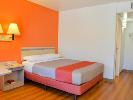 A simple hotel room with a bed, orange accent wall, wall-mounted lamps, bedside table, and open closet area in a wood-floored space.