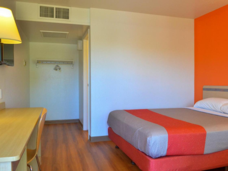 A hotel room with a bed, desk, wall lamp, TV, and open closet. The decor is simple with an orange accent wall and wood flooring.