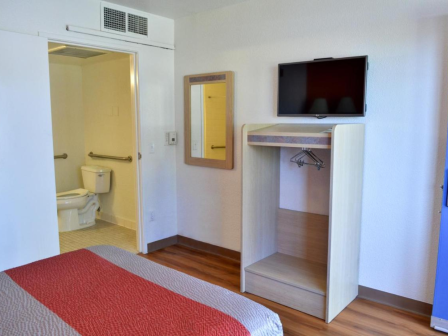 A hotel room with a bed, wall-mounted TV, open closet, mirror, and attached bathroom with visible toilet and grab bars.