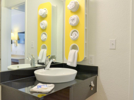 A bathroom with a sink, wall-mounted mirror, faucet, towel holder with rolled towels, and a small countertop with a soap packet.