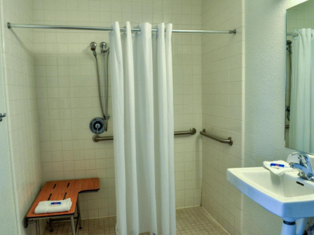 The image shows an accessible bathroom with a shower area, curtain, bench, grab bars, sink, and a mirror.