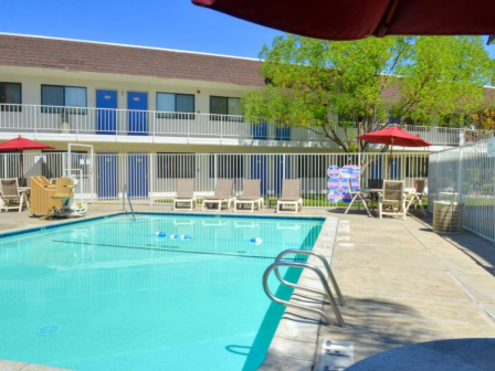 A clear swimming pool with a metal railing, surrounded by lounge chairs and umbrellas, near a two-story building with blue doors.